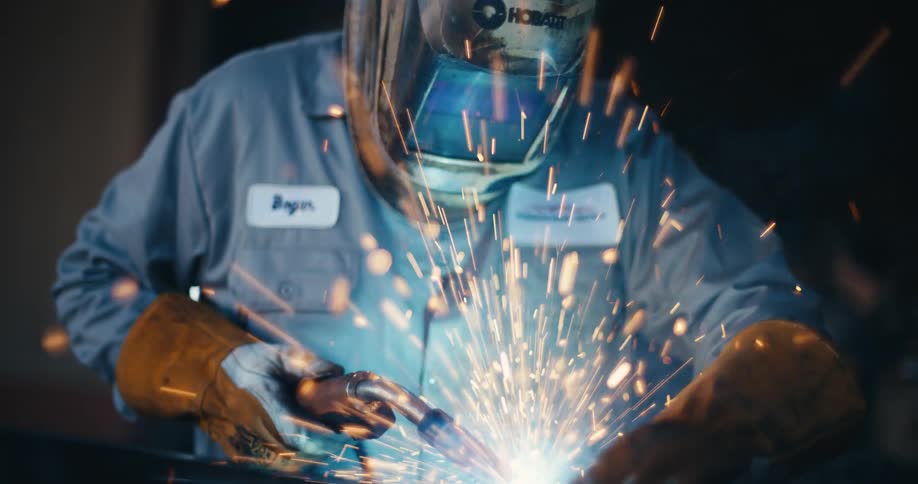 An industrial welder wearing a protective mask works on a piece of metal, creating a brilliant shower of sparks in slow motion