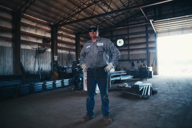 A proud and confident blue-collar worker stands with his hands on his hips in a large industrial metal shop as the camera slowly zooms out