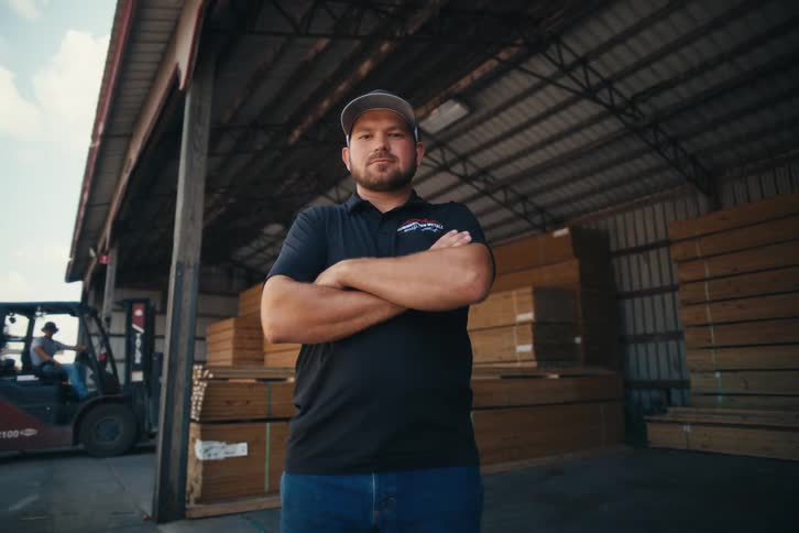 A confident male worker with his arms crossed stands inside a lumber warehouse with a forklift in the background