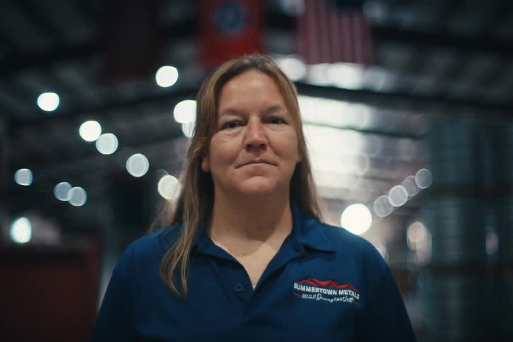 A confident female factory worker turns to look at the camera inside a large industrial warehouse