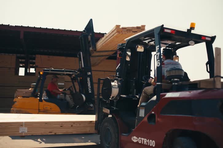 Two workers operate forklifts to move and stack lumber at an outdoor supply depot on a sunny day