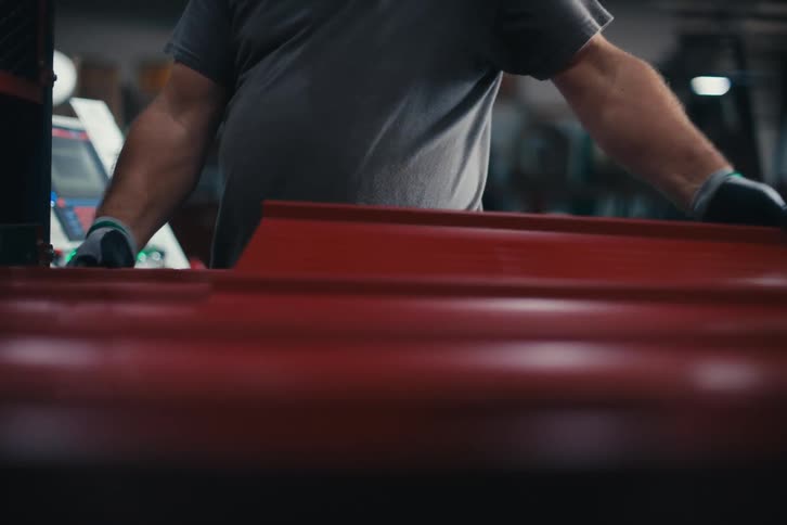 A factory worker with ear protection carefully oversees the production of red corrugated metal roofing sheets on a machine