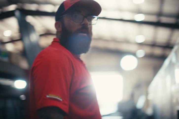 A factory worker with tattoos and a beard carries a large sheet of metal through a warehouse in slow motion