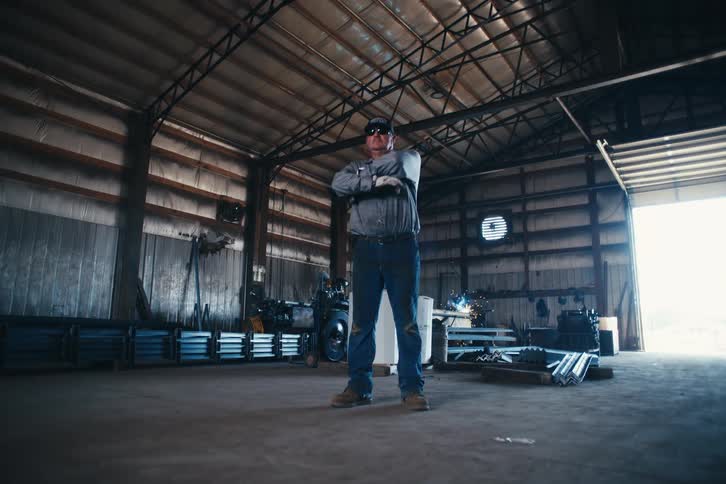 A confident blue-collar worker proudly crosses his arms while standing inside a large industrial metal workshop