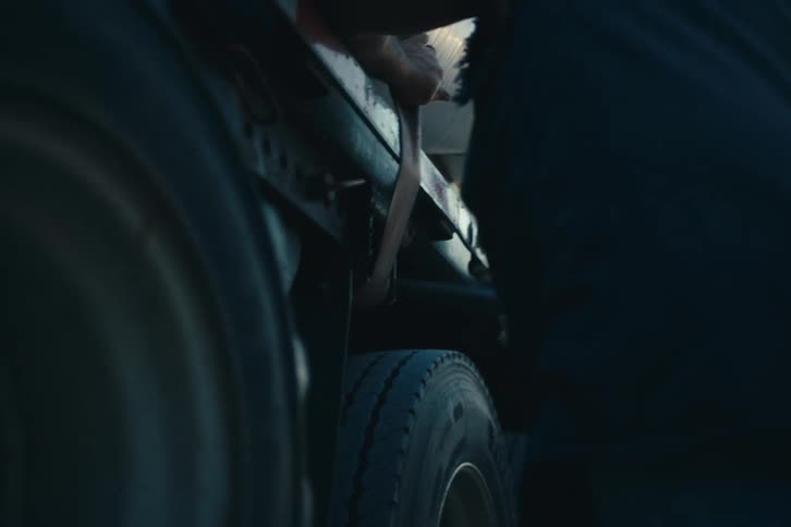 A truck driver uses a winch bar to tighten a ratchet strap securing large white bundles on a flatbed trailer