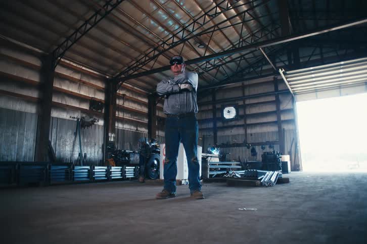 A proud welder stands with arms crossed in his workshop as sparks fly from welding in the background