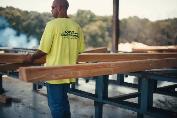 Two construction workers at a lumberyard cut and move large wooden beams on a sunny day