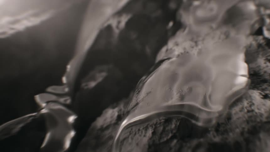 A viscous, clear liquid flows in slow motion over a dark, textured, rocky surface in this abstract macro shot