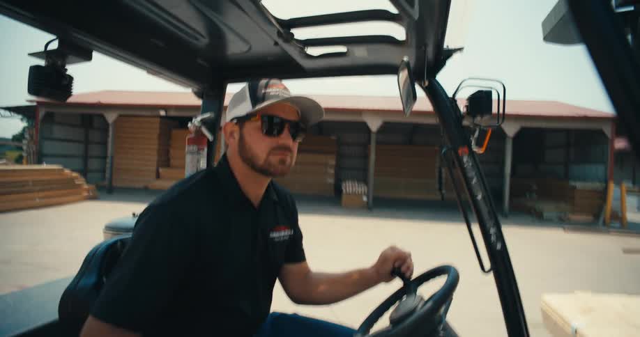 A forklift operator in a cap and sunglasses drives through an industrial lumberyard on a sunny day