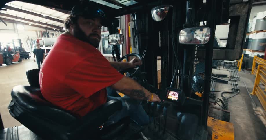 A male forklift operator in a red shirt drives a forklift in reverse through an industrial warehouse