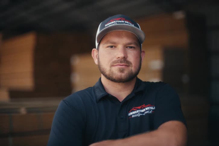 A confident, bearded worker in a company uniform and cap turns to look at the camera and crosses his arms in a warehouse