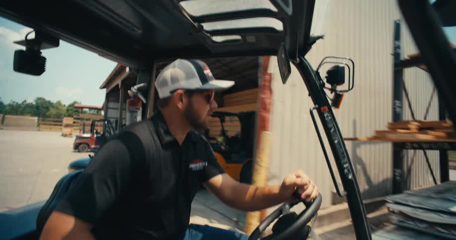 A man in a hat and sunglasses drives a forklift through a busy industrial lumberyard on a sunny day