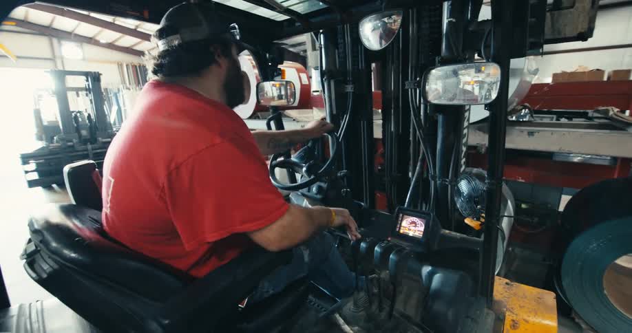 A bearded man in a red shirt drives a forklift through a manufacturing warehouse filled with materials