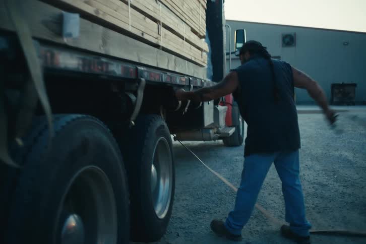 A male truck driver pulls on a tie-down strap to secure a load of lumber onto his flatbed truck in a yard at dusk