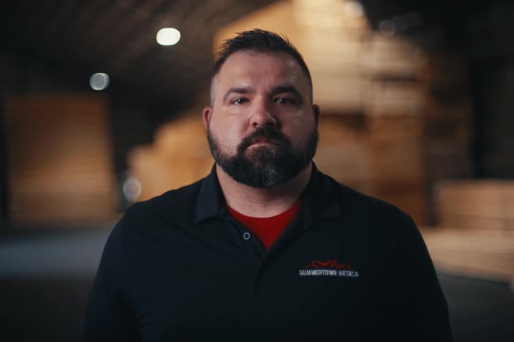 A confident, bearded man stands stoically inside a dimly lit lumber warehouse