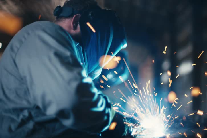 A skilled metalworker in a protective mask performs arc welding on a steel structure, creating a brilliant shower of sparks and smoke in a dark workshop