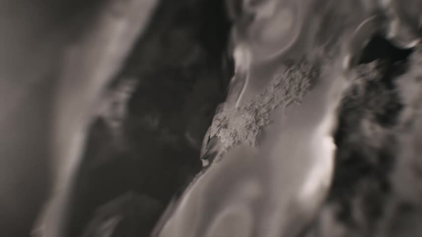 A clear, viscous fluid slowly seeps over a dark, textured surface in a dramatic black and white macro shot