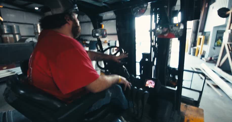 Bearded warehouse worker in a red shirt drives a forklift forward towards the bright light of an open bay door