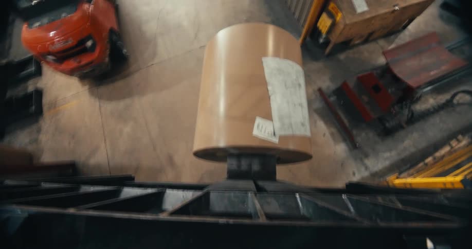 From a high angle perspective, a large industrial roll of metal is being transported on a machine through a factory warehouse