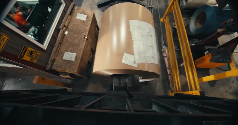From above, a large industrial machine unwinds a massive coil of sheet metal in a factory setting
