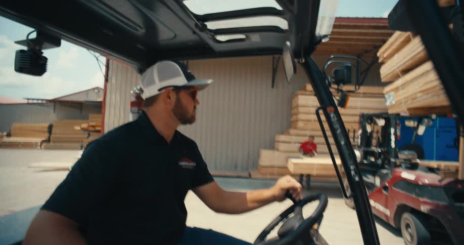 A forklift operator in a cap and sunglasses watches another worker move lumber at a supply yard