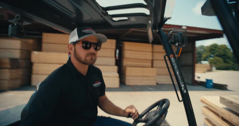 A confident worker in sunglasses and a hat drives a forklift through an industrial lumberyard on a sunny day