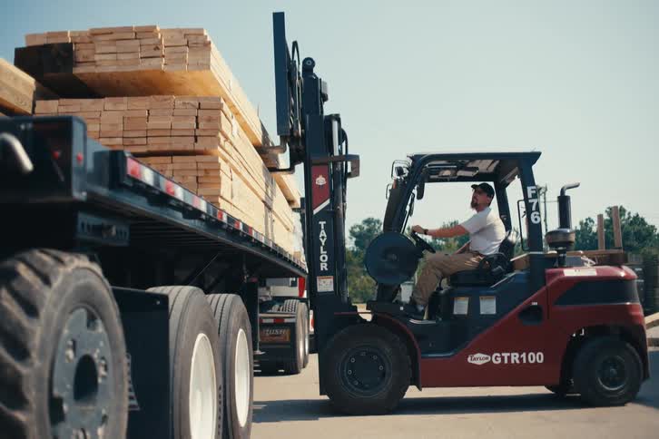 A forklift operator carefully loads stacks of lumber onto the flatbed of a large truck at an industrial yard on a sunny day