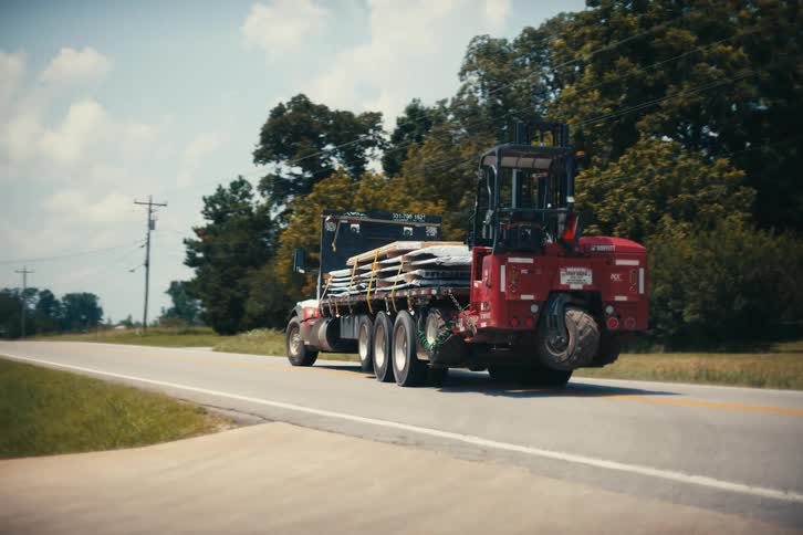 A flatbed truck with an attached forklift drives away down a rural road carrying a load of materials