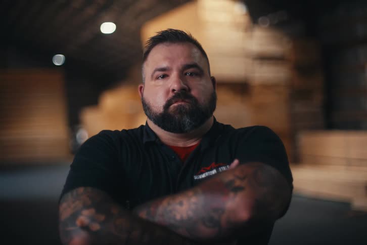 Portrait of a strong, tattooed worker with a beard, crossing his arms and looking seriously at the camera in a lumber warehouse