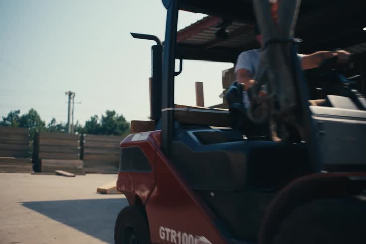 A man operates a red forklift, moving building materials at an outdoor lumber yard on a sunny day