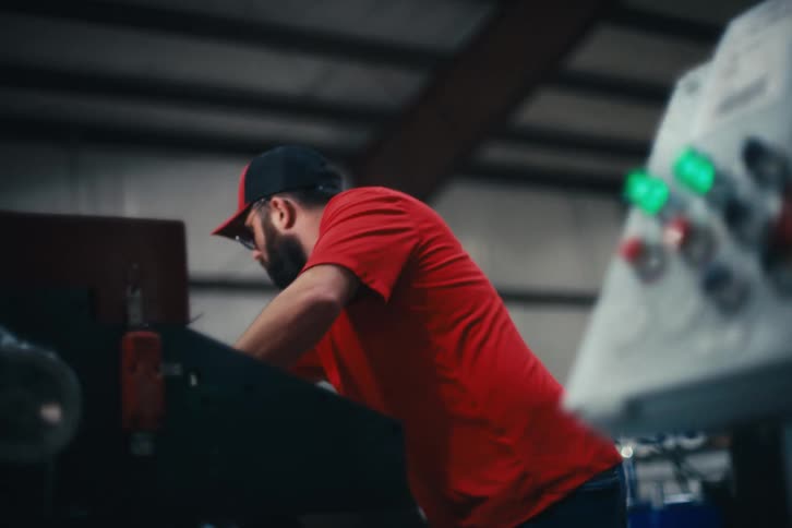 A man with a beard and glasses works on a machine in a manufacturing facility