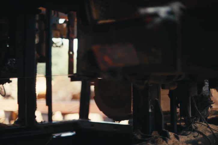 A large bandsaw slices through a round wooden log in a dusty, dimly lit sawmill