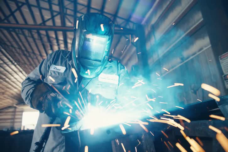A welder in a protective mask works on a metal piece in a workshop, creating a shower of bright sparks in slow motion