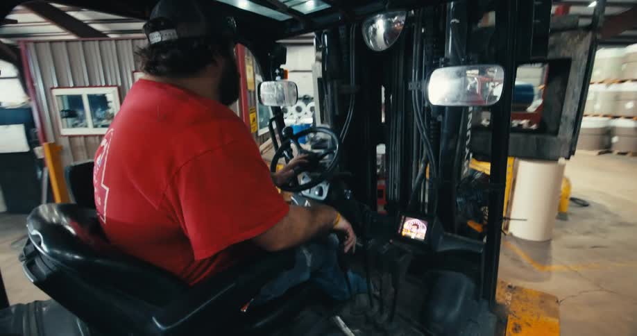 A bearded man in a red shirt and baseball cap operates a forklift, carefully driving through an industrial warehouse