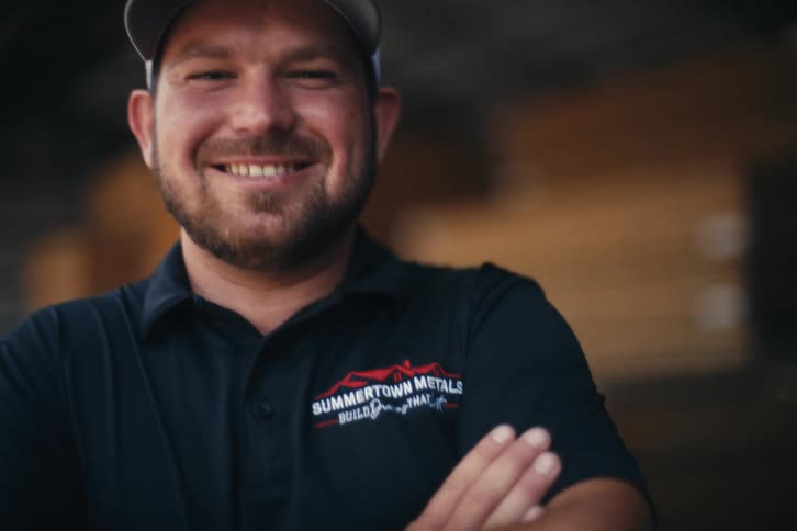 A friendly, bearded construction worker in a branded hat and polo shirt smiles confidently at the camera