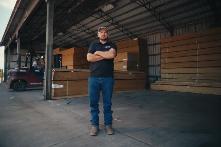 A confident male worker stands with arms crossed in a lumber warehouse as a forklift operates in the background