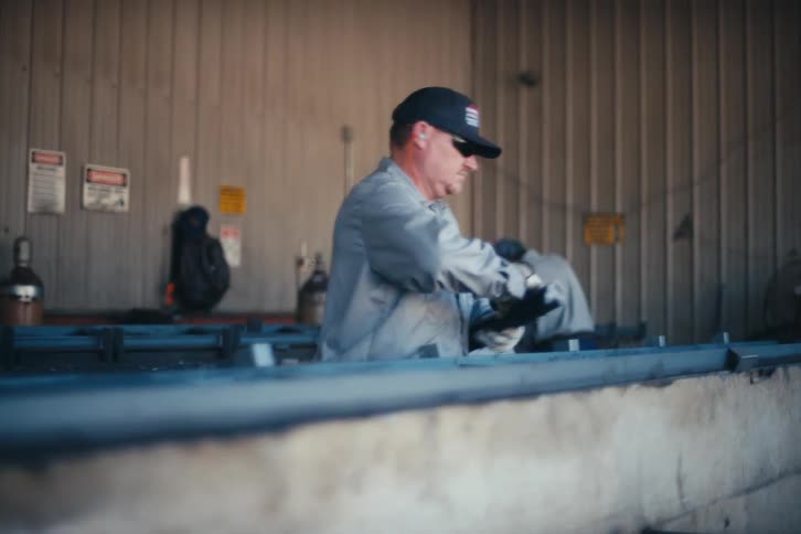 A metalworker assembles a large steel frame while colleagues weld in the background of an industrial factory