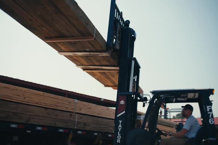 A low-angle view shows a forklift operator skillfully loading large wooden beams onto a flatbed truck