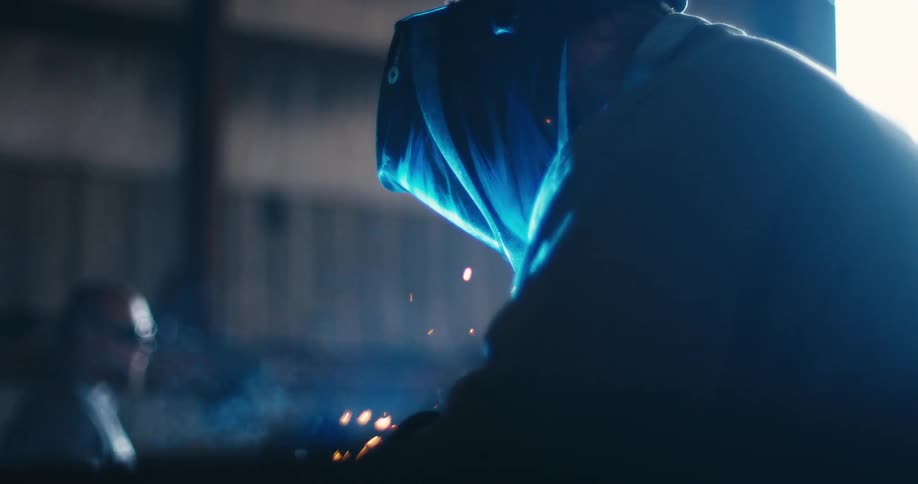 Slow motion close up of a welder's helmet as bright blue light and orange sparks fly from the tool