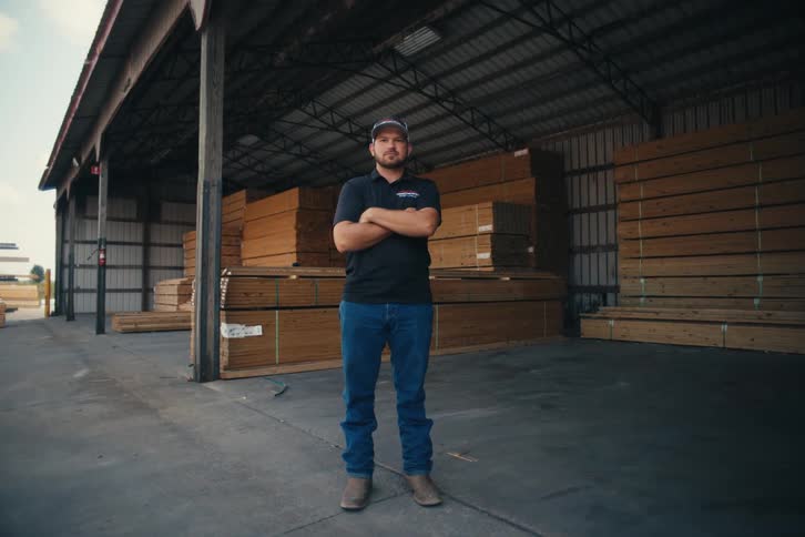 A male lumber yard worker in a baseball cap confidently crosses his arms in a warehouse full of stacked wood