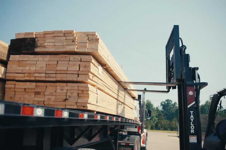A forklift slowly backs away from a flatbed truck fully loaded with stacks of lumber at a supply yard