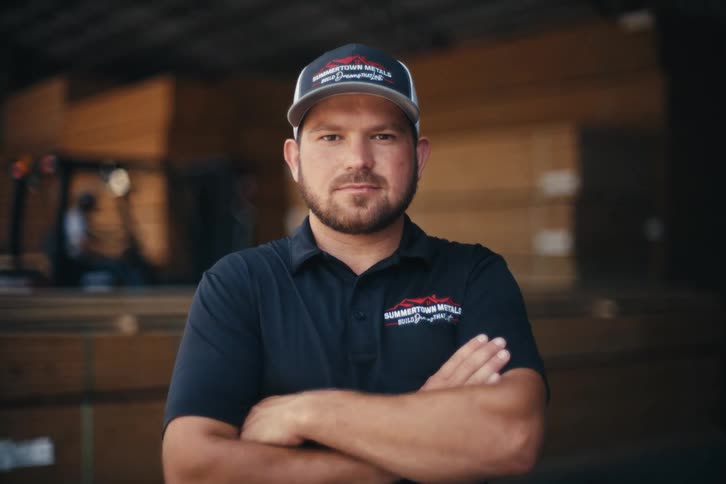 A male worker with a beard and hat crosses his arms and looks confidently at the camera in a lumber warehouse