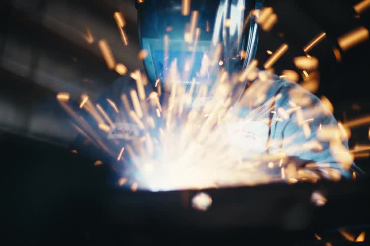 A welder in a protective mask works on a piece of metal, creating a shower of bright sparks in a workshop