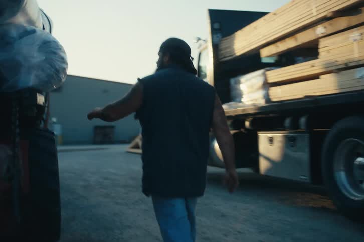 A male truck driver walks to his flatbed trailer and tightens the straps to secure the cargo in slow motion