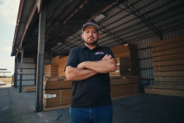 A confident male worker in a baseball cap crosses his arms while standing inside a large lumberyard warehouse
