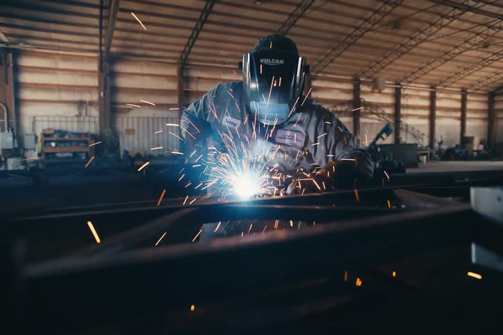 A slow pan reveals a welder in a protective mask working diligently on a metal beam inside a large, industrial warehouse