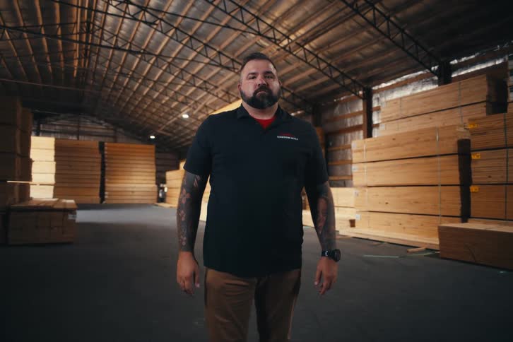 A confident, tattooed man stands seriously looking at the camera inside a large lumber warehouse