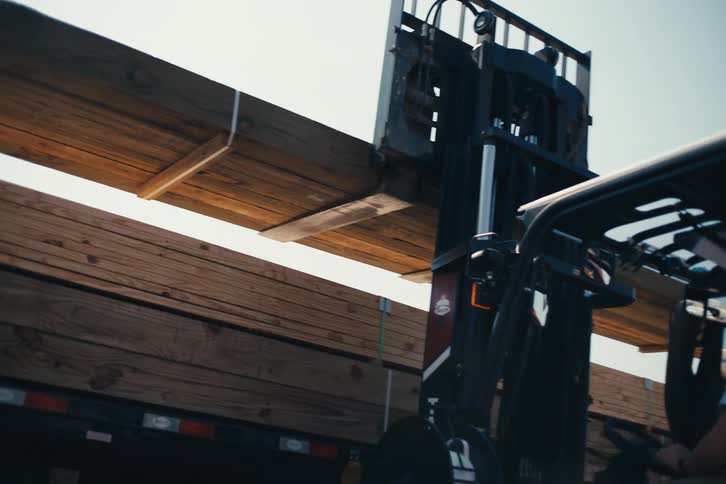 A forklift operator carefully loads a large bundle of lumber onto a flatbed truck in slow motion