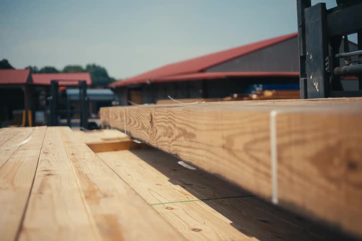 A forklift carefully lowers a stack of fresh wooden planks onto another stack in a sunny lumber yard