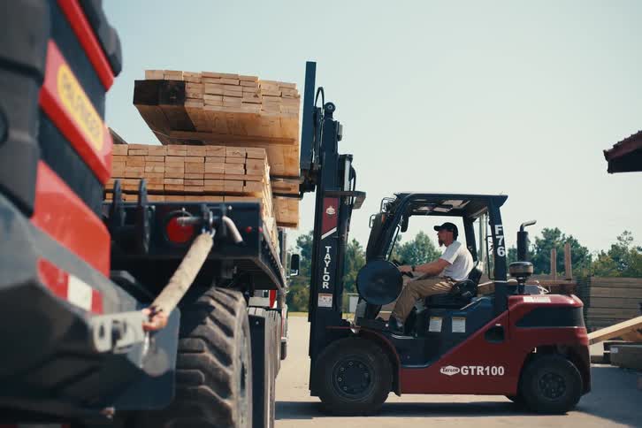 Forklift operator carefully loads a stack of lumber onto a flatbed truck at a sunny industrial yard